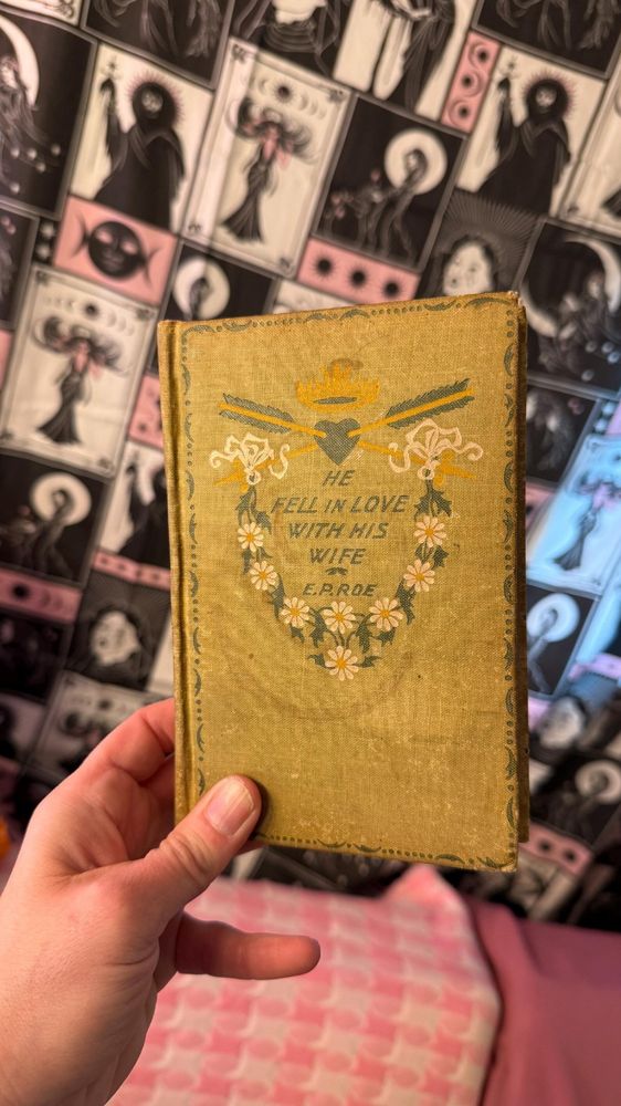 holding up a hand-bound journal with an old book cover. the book is titled "He Fell in Love with His Wife" by E.P. Roe.