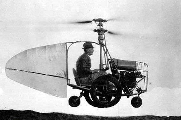 Svartvit historisk bild som visar en man i en tidig experimentell flygfarkost. Farkosten har en stor propeller ovanpå, liknande en helikopterrotor, ett enkelt säte och en ram som påminner om en cykel eller ett litet fordon med tre hjul. Till vänster sitter en vingliknande konstruktion som fungerar som stabilisator eller lyftyta. Mannen bär hatt och sitter upprätt medan fordonet svävar strax ovanför marken mot en ljus himmel i bakgrunden.