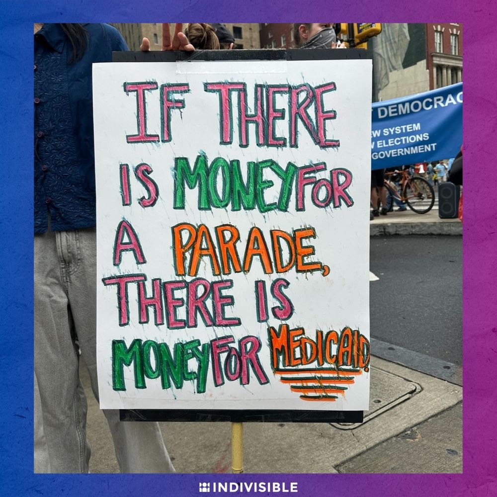 A protest sign reads "If there is money for a parade, there is money for medical" in colorful letters at a demonstration. The "Indivisible" logo is at the bottom right corner.