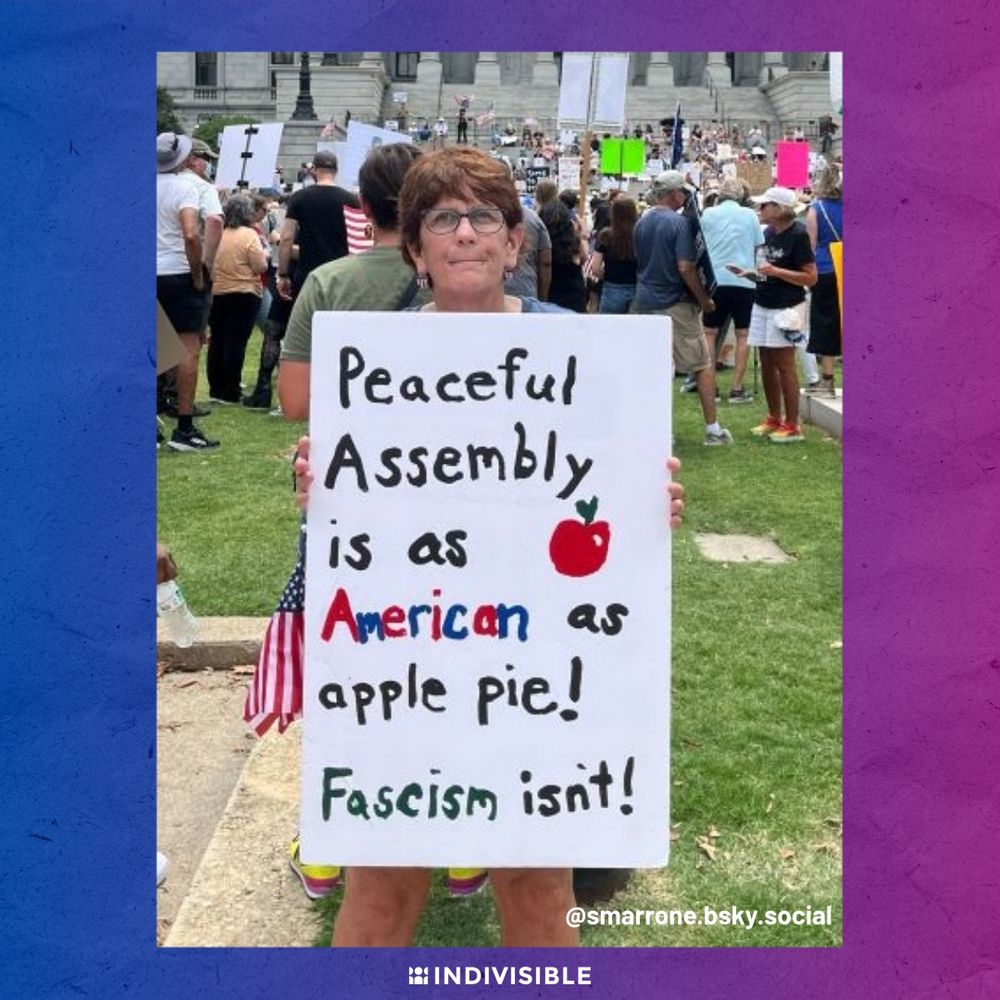 Person holding a sign at a protest that reads "Peaceful Assembly is as American as apple pie! Fascism isn't!" in front of a large crowd in front of a government building.