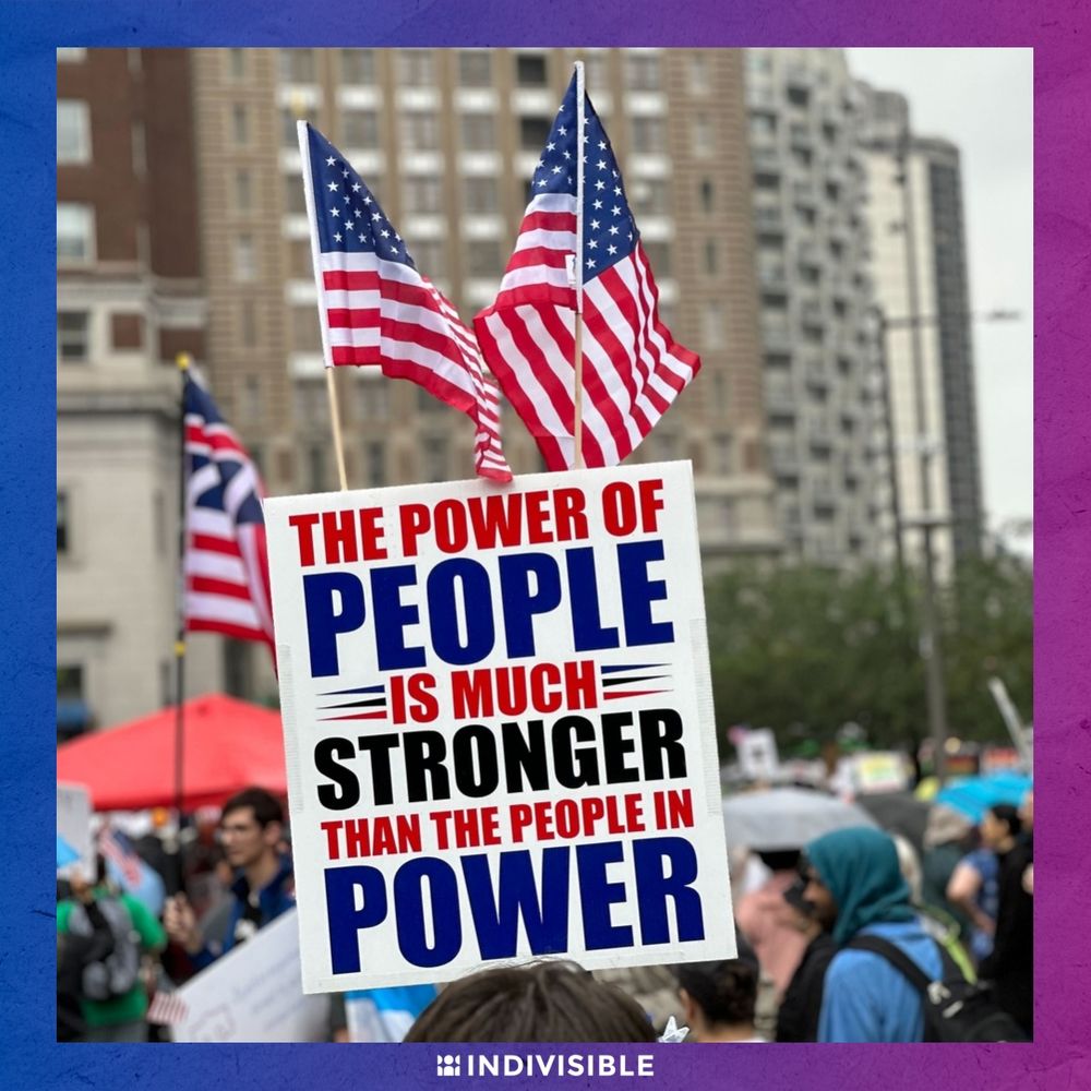 A protester holds a sign reading "The power of people is much stronger than the people in power" with two American flags in the background, at an Indivisible rally.