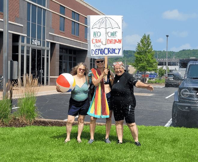 Three Wisconsin Indivisibles in bathing suits holding a towel, a beach ball, and a sign that reads “NO ONE CAN DROWN OUR DEMOCRACY.”