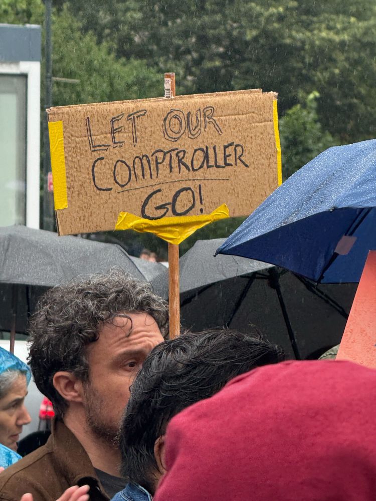 A protester holds a sign that reads “LET OUR COMPTROLLER GO!”