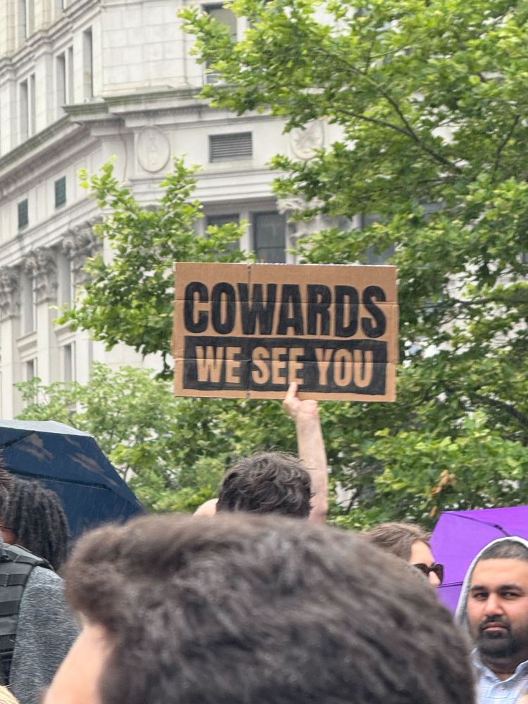 A protester holds a sign that reads “COWARDS/WE SEE YOU.”
