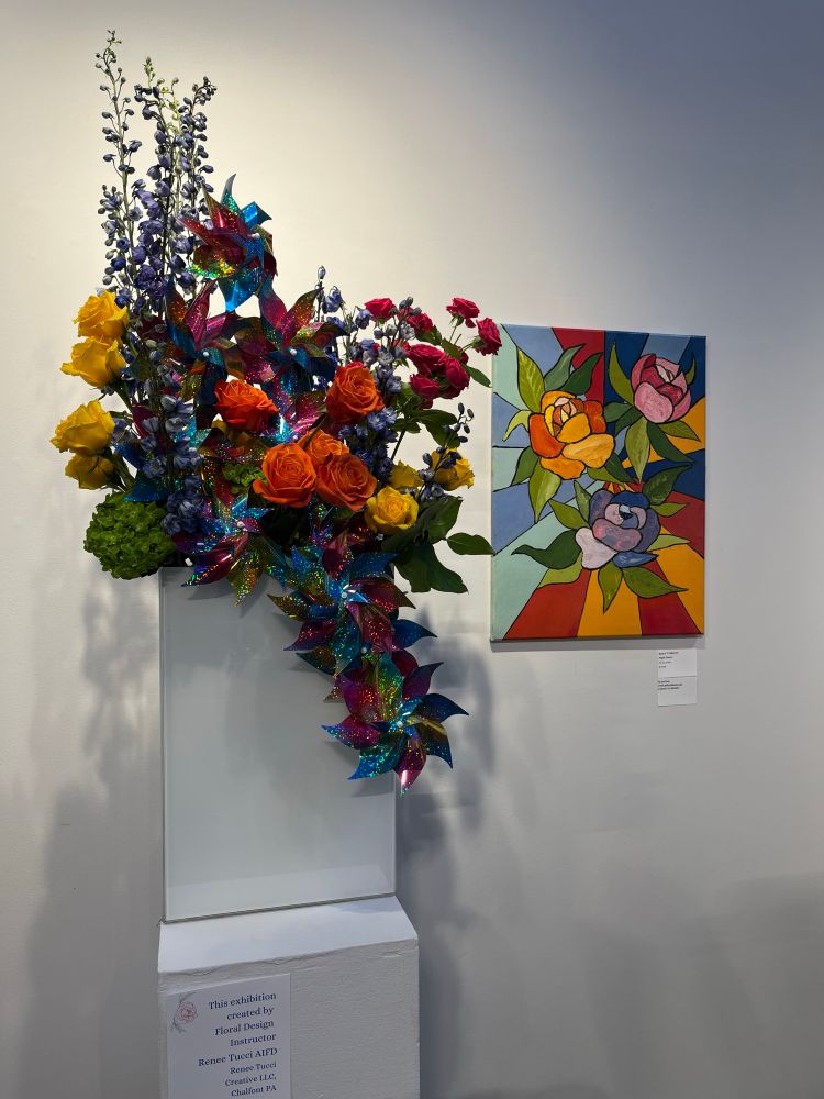 The acrylic painting on the wall depicts three abstracted cabbage roses using a boldly fauvist palette on a graphic background dominated by vividly colored rays emerging from a central point. The oversized bouquet echoes the painting’s palette with deep yellow, orange, and red roses, eucalyptus sprigs, and a cascade of glittery plastic pinwheels. 