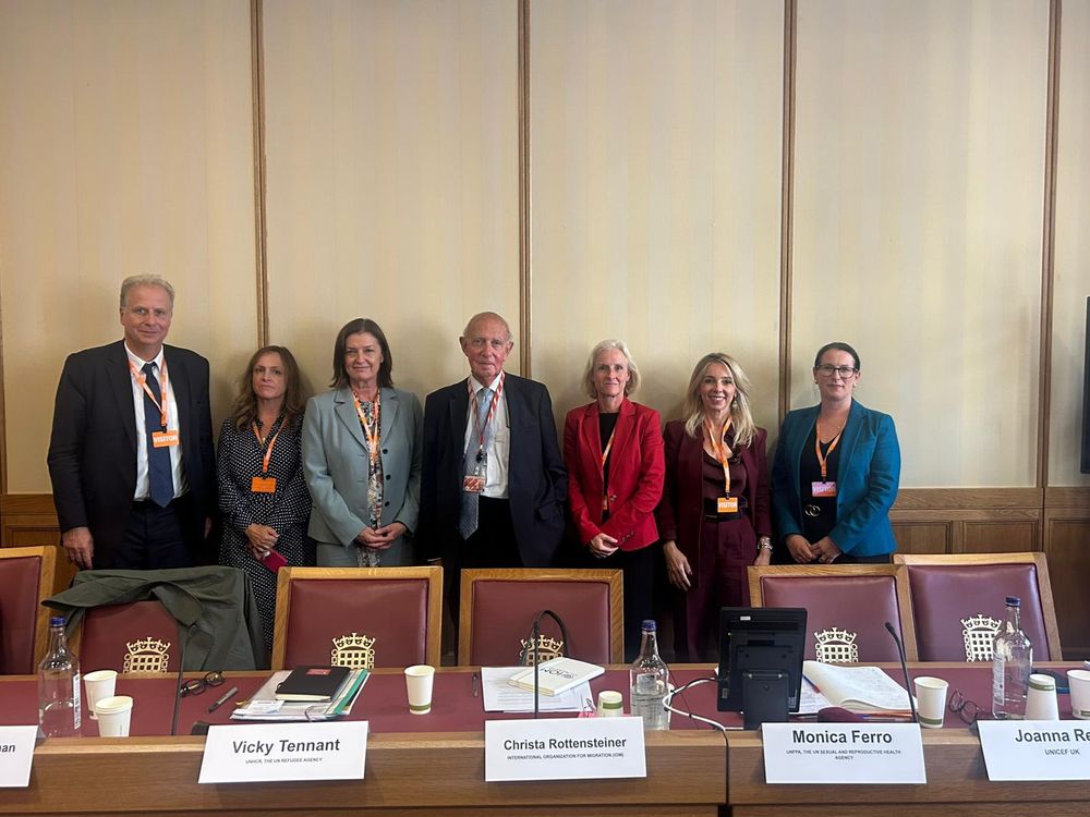 7 people stand infront of a desk and chairs in a formal setting in UK parliament 