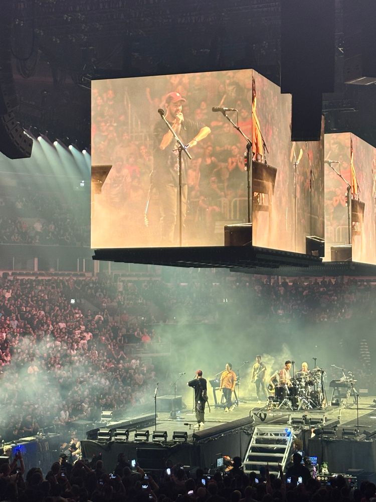 Closer view of the stage, with more light present while Mike Shinoda raps to the audience. He is also viewable on the large overhead screens.