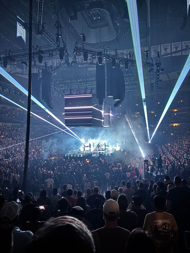 Mid range view of an arena stage surrounded by fans, with two huge cube-shaped screens hanging above and bright white spotlights aimed in various directions. Linkin Park is silhouetted in white light on the stage as they play.