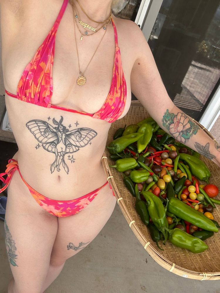 the bikini gardener posing with a large hip winnower filled with vegetables 