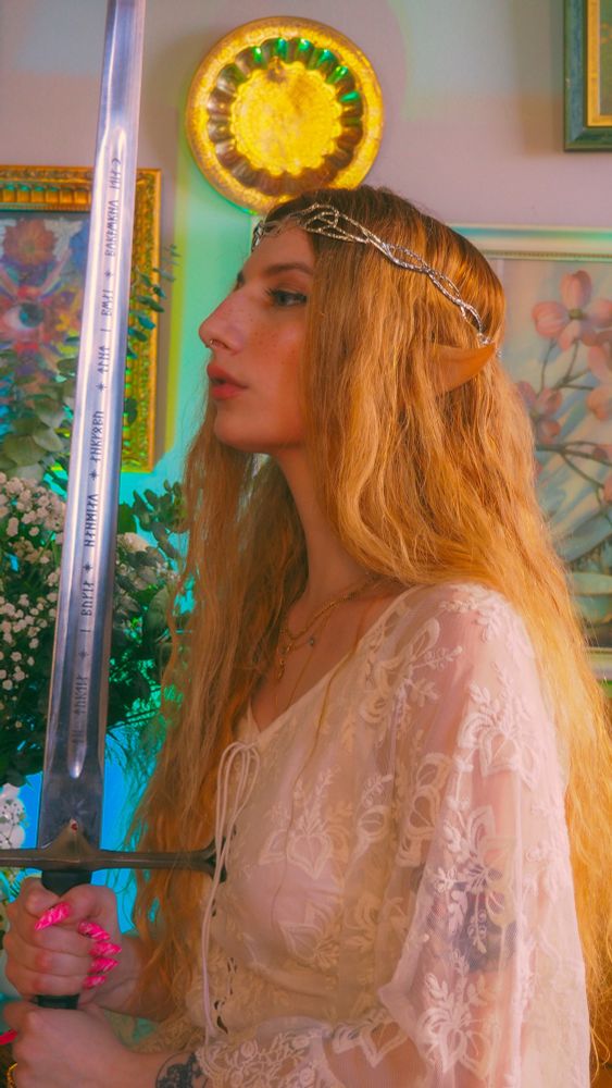 marina at a full profile dressed as galadriel in all white. they’re holding the sword anduril up to their face as if ready to knight someone