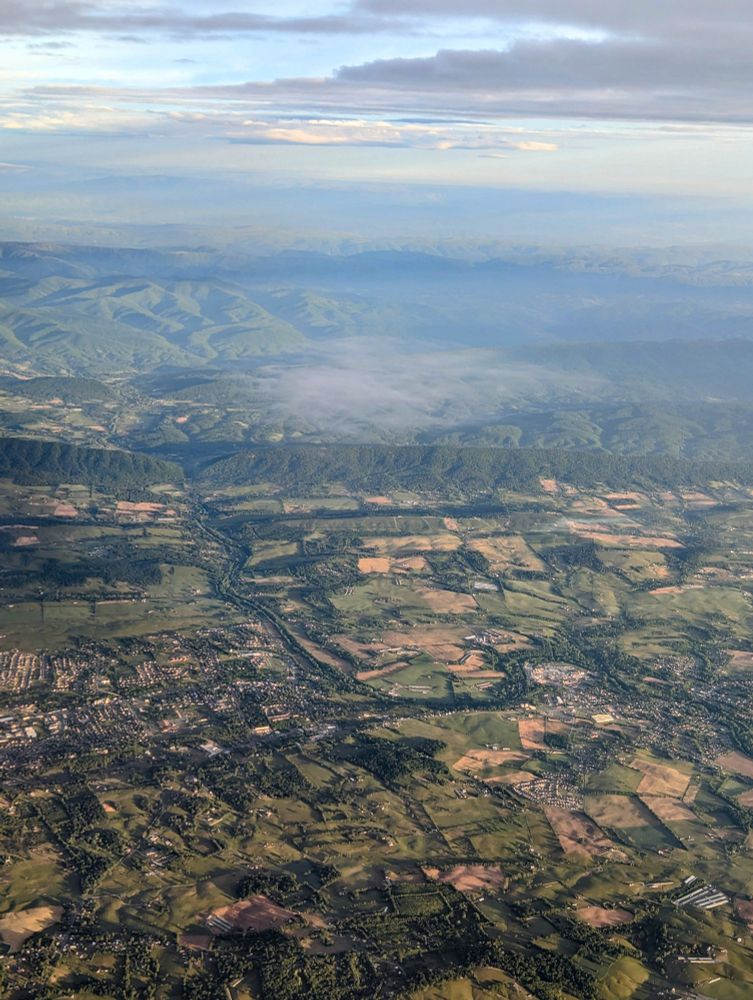 View from an airplane flying over part of Virginia with forested mountain ridges on the edge of a valley with small farms, houses, and a river that passed through the mountains 