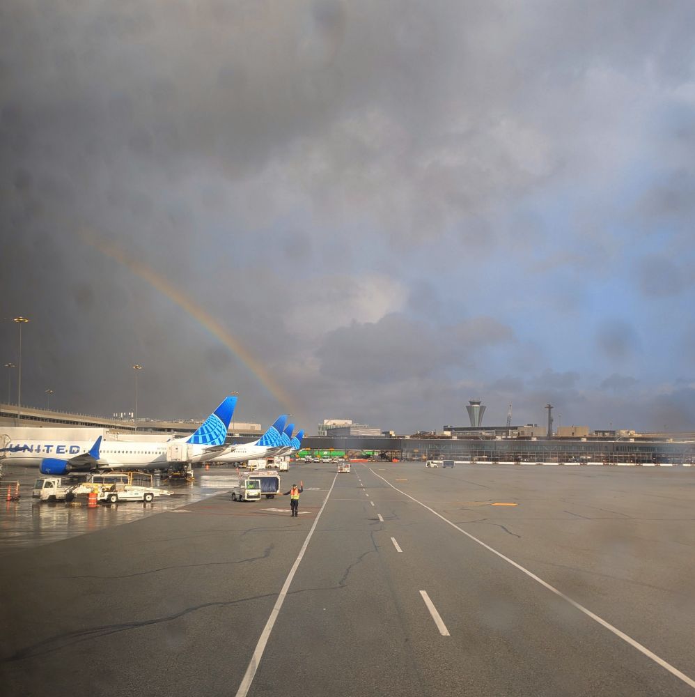 View from a plane pulling into the gate of an airport with a rainbow and mostly cloudy sky in the background. There are raindrops on the window.