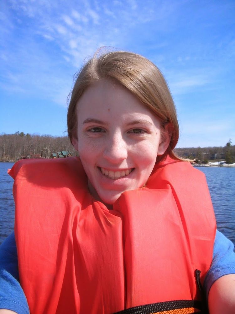 Lindsay, much younger, wearing a bright red life vest. She's taking a selfie. She has light brown hair and has a big smile