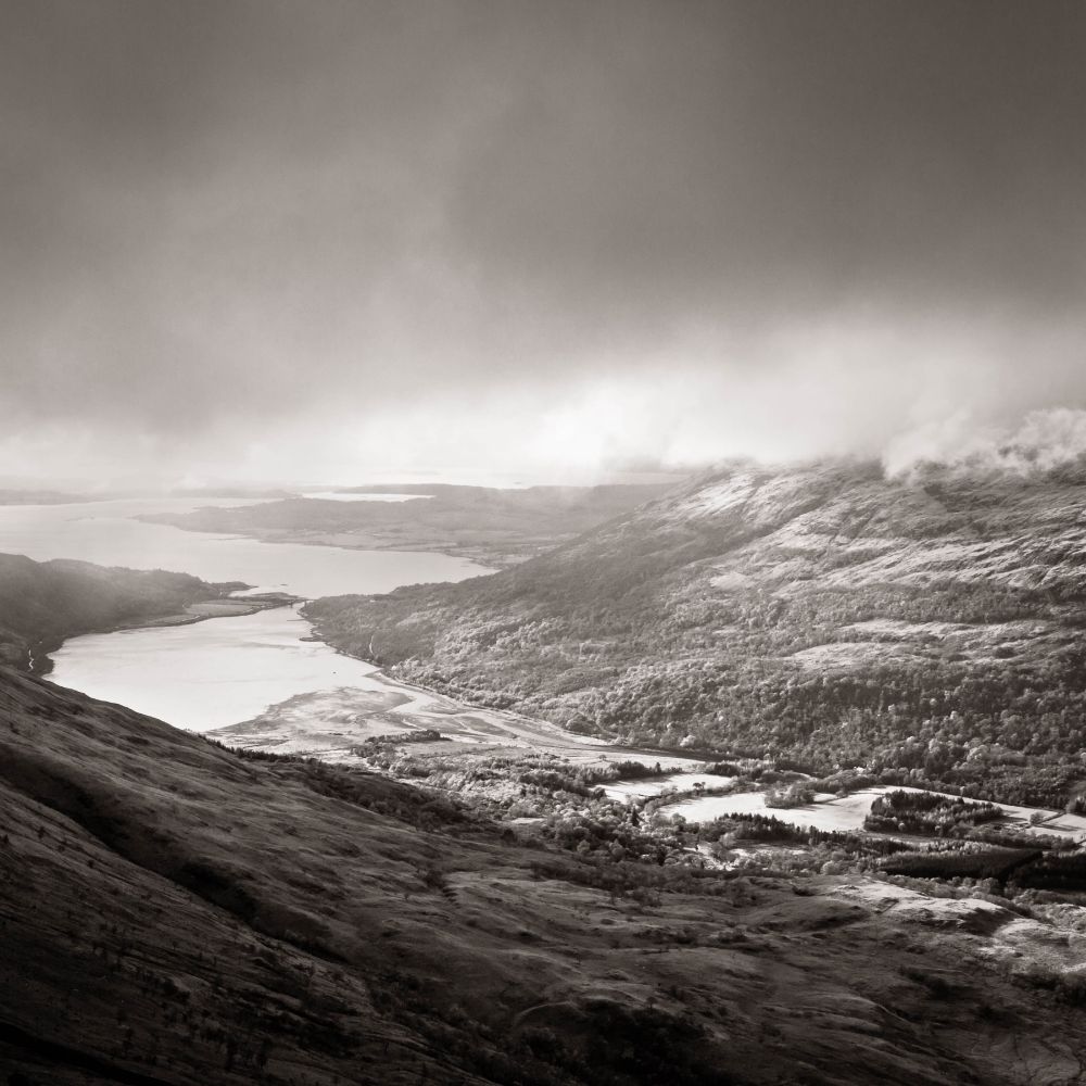 A black-and-white image taken from a Scottish mountain. After a dull day, late afternoon sunshine finally breaks through the clouds and illuminates the glen and loch below. Scotland never disappoints! 