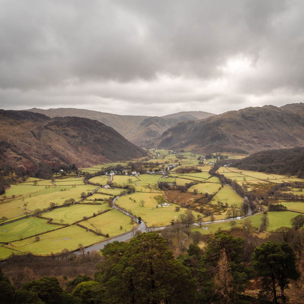 A view over Borrowdale as seen from Castle Crag in the Lake District. A patchwork of green fields cover the valley floor, surrounded by rugged fells. A grey day when the greens look vibrant.