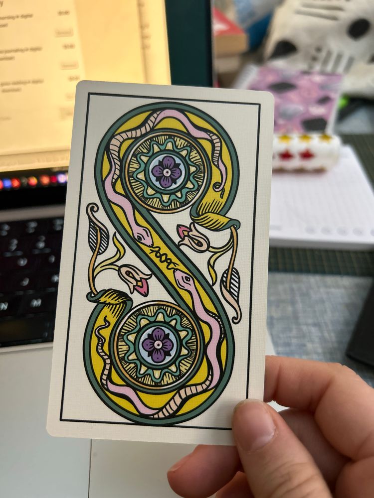 A hand holding up the 2 of Disks card from the Gay Marseilles tarot. The card looks like a big S with disks in the curves. It is illustrated in an updated Marseilles style, with two snakes facing each other in the S itself. There are flowers and other flourishes. The color scheme is a canary yellow, light pink, teal, green, sky blue, and purple