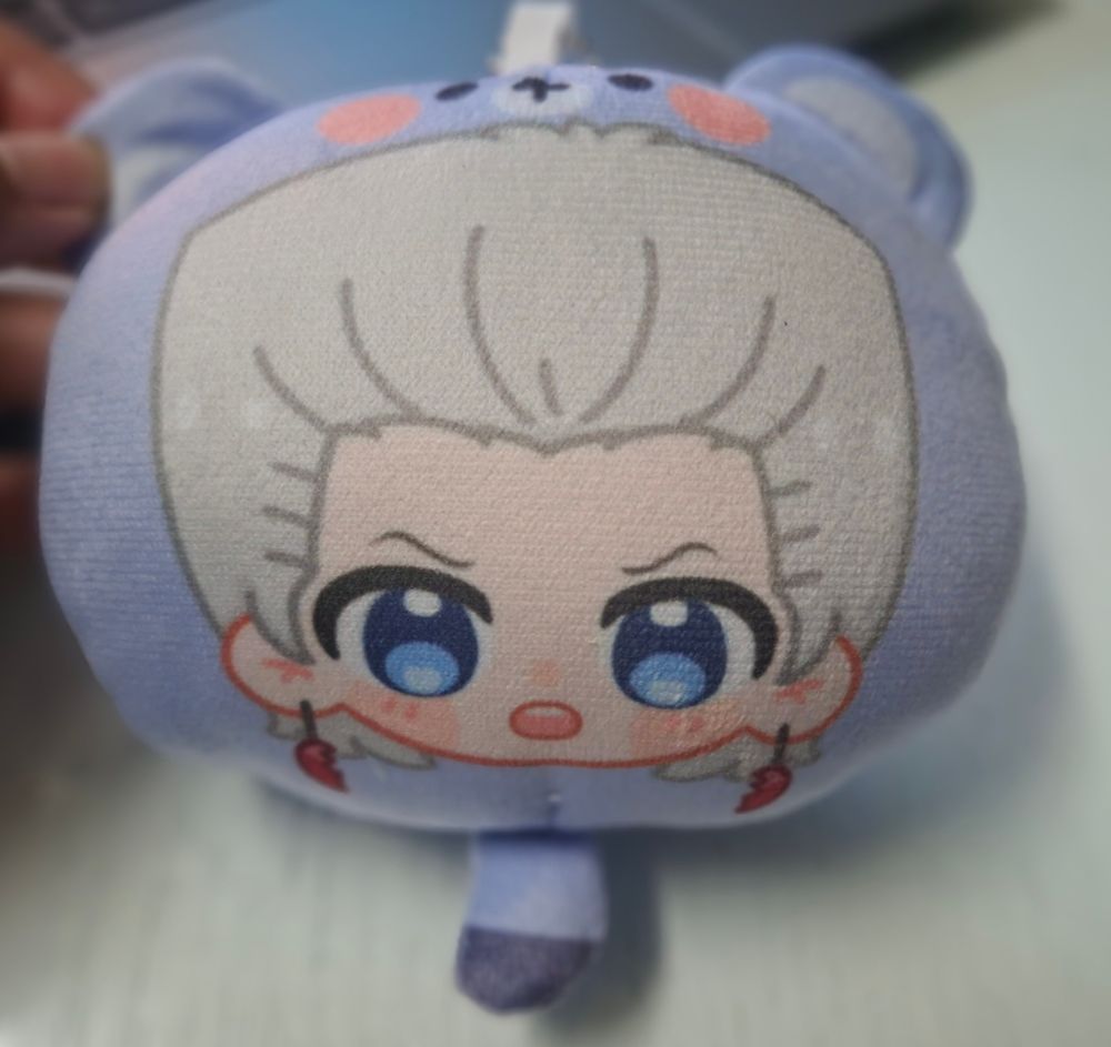 Plush keychain of a purple bear/racoon(?) with a fanart of chibi Polnareff printed on it.