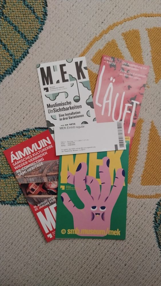 Eine Collage von Museumsheftchen über einzelne Ausstellungen im MEK (Museum Europäischer Kulturen) in Berlin, darauf liegt eine Eintrittskarte mit dem heutigen Datum 17.11.24 