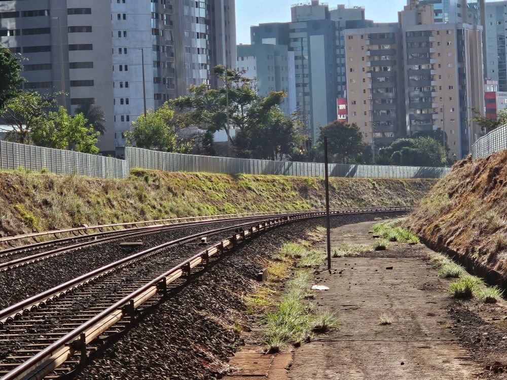 Foto da luz da manhã nos Trilhos do metrô da estação Arniqueiras em Brasília - DF. Nesse ponto o trem circula na superfície então da pra ver o gramado e algumas árvores e edifícios atrás das cercas