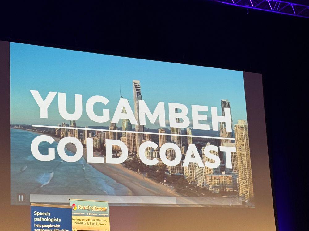 A large screen displays an aerial view of the Gold Coast skyline with the words “YUGAMBEH GOLD COAST” overlaid in bold white text.