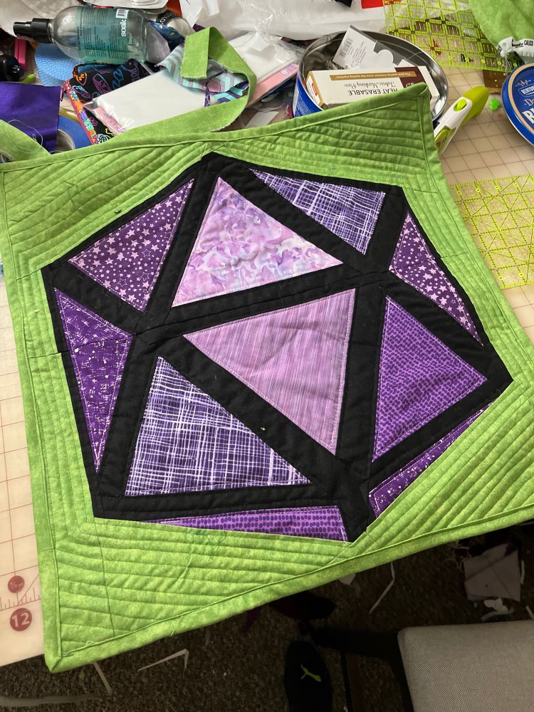 A mini quilted wall hanging featuring a purple 20-sided die with a green background.
