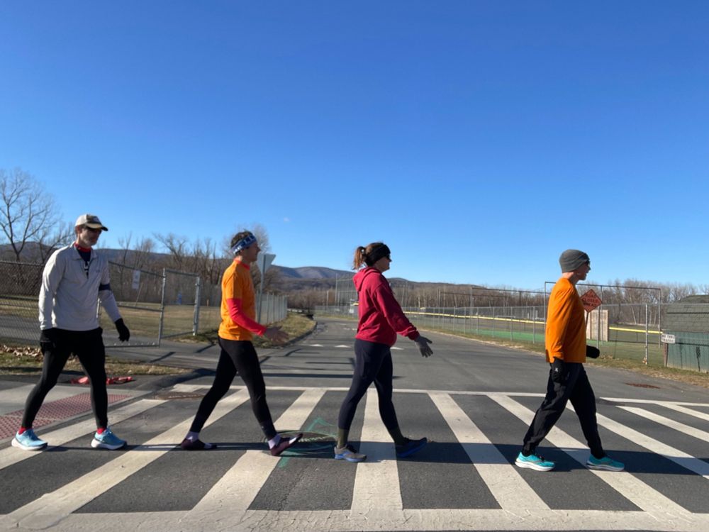 Four runners crossing a street at a crosswalk, looking astonishingly like the Beatles. The sky is blue and it is cold out.
