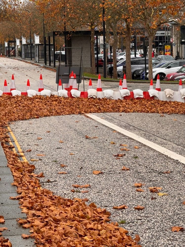 A twin carriageway in Milton Keynes, parked cars in the background. The road is blocked by cones, barriers, and sandbags, and brown fallen leaves have gathered up against the barrier
