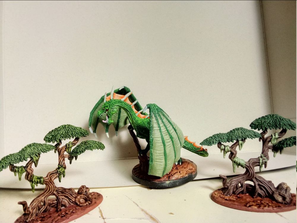 The miniature of a green dragon perching on a tree stump, peeking out from behind its wing, side view. Left and right are mangrove-style tree-miniatures