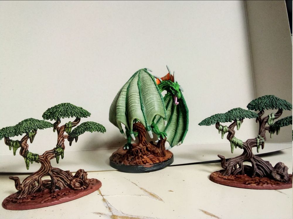 The miniature of a green dragon perching on a tree stump, peeking out from behind its wing. Left and right are mangrove-style tree-miniatures