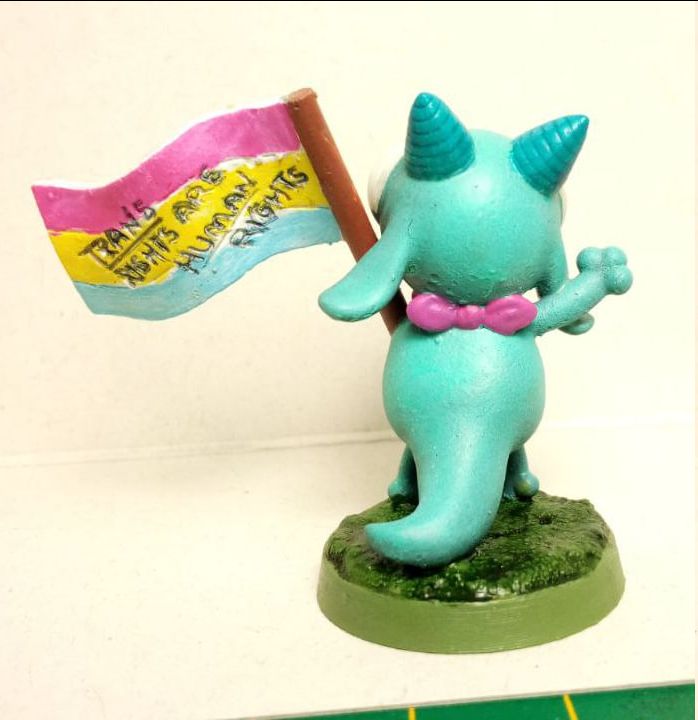 The backside of the turquoise kobold in a cartoony style with pink bow waving a pan pride flag. Trans Rights Are Human Rights is written on the flag. Miniature is a 3D print I ordered from mini-marvels.nl, and comes from Found Family Adventurecrafts!