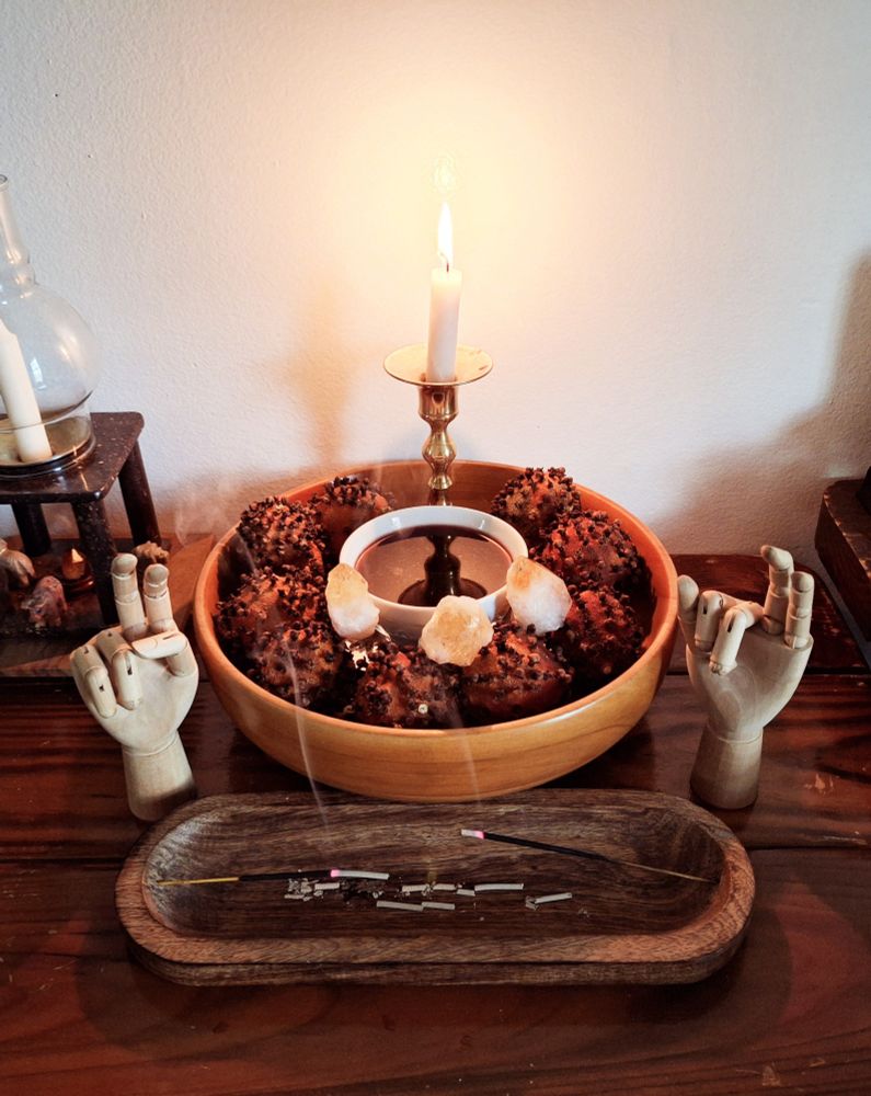 A white candle burning in a wooden bowl filled with dried pomanders surrounding a small bowl of wine. Incense burns in front in a wooden holder. Two small wooden hands make sacred gestures on either side.