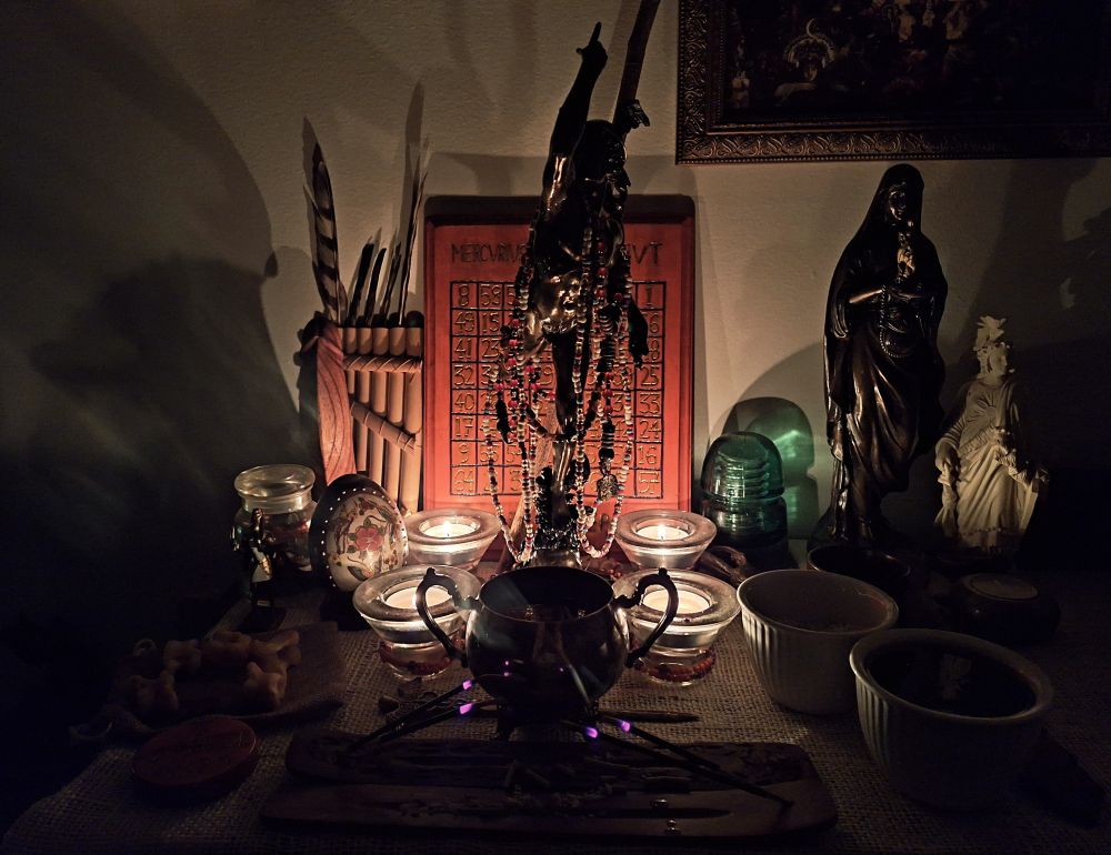 A dimly-lit modern shrine of Hermēs, with a statue of the god surrounded by four tealights lit and sticks of incense burning in front with various offerings and other shrine paraphernalia about.