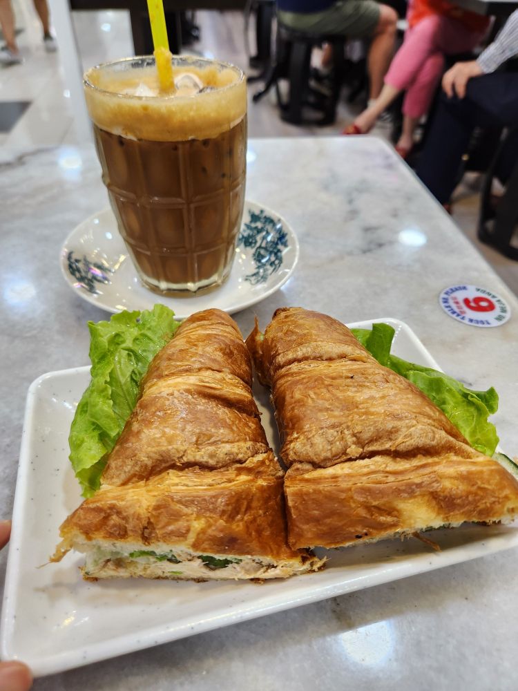 A croissant served on a plate, cut in half, filled with tuna mayo, sliced cucumbers, and fresh lettuce. Beside it is a cup of iced "cham" served on tiny dish. "Cham" is Cantonese for "Mixed" tea and coffee.