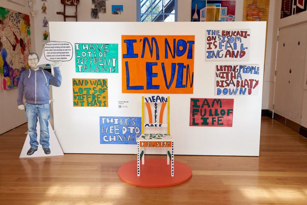 Photo of an exhibition. A white wall features 7 bright paintings with a chair placed in front of it. The main painting in blue and orange reads “I’m not leaving”
