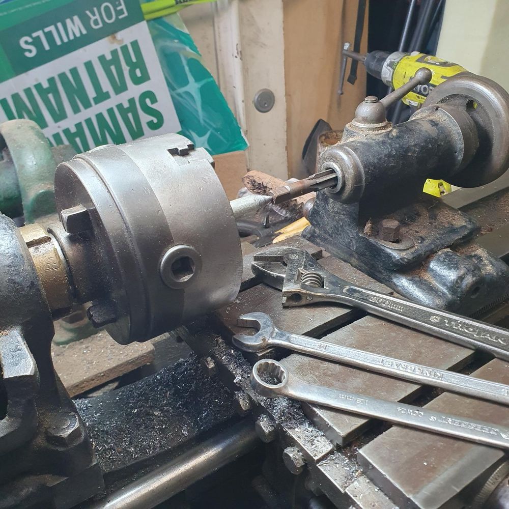 the chuck of a Drummond M-Type lathe with a dead center (pointy bit of steel) in it, supporting a reamer which is inserted into the tailstock. Everything is greasy and covered in metal filings, and there are an assortment of spanners on the crossbed