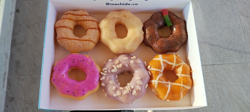 Box of 6 mochi doughnuts. From top left clockwise: churro, salted butter, dirt pie, white chocolate pumpkin, taro crisp, and dragon fruit.