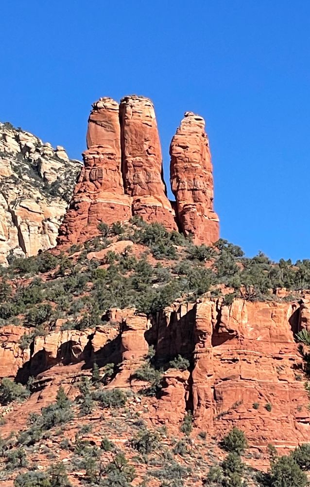 Three finger-like stone pillars jut up from a hill above redrock cliffs.  In the background a buff-colored mountain rises to the left, all beneath clear blue sky.