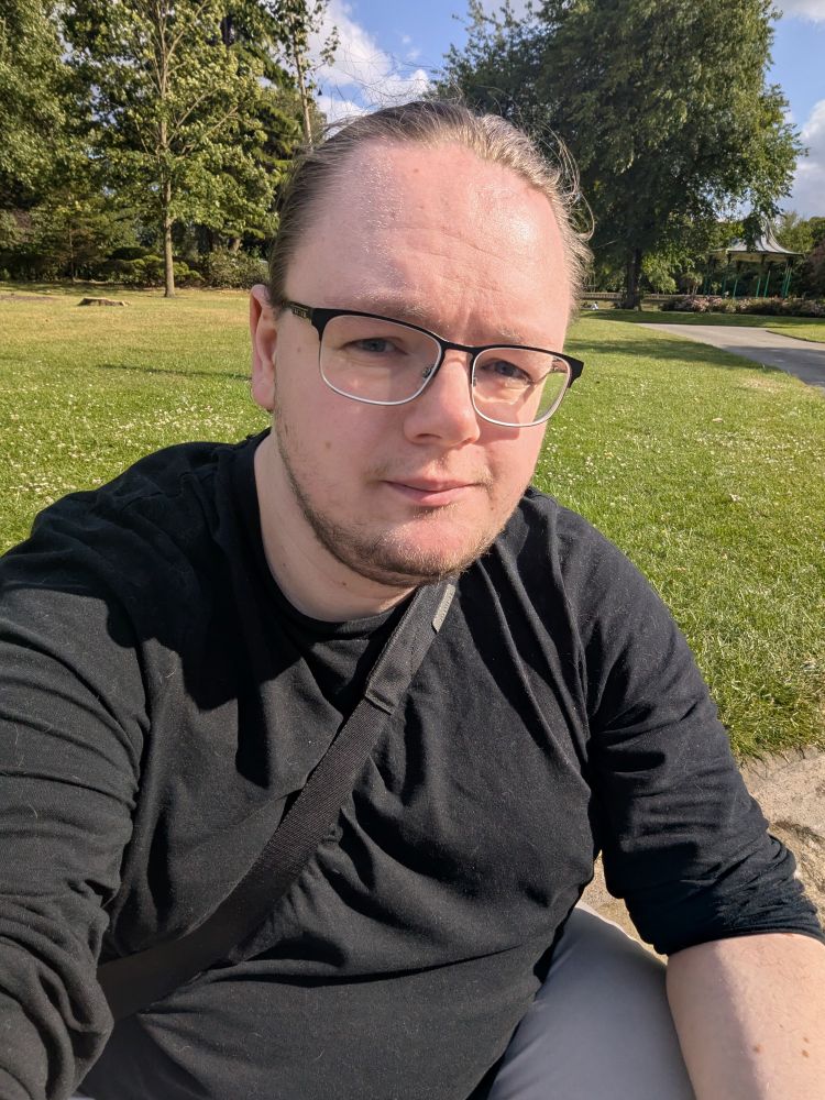 an image of me, a white masc-presenting non-binary person with tied back hair and new, kinda round glasses, wearing a black long-sleeved shirt and a bag over my chest. my posture is very poor and my eyes are squinting because it's very sunny. 