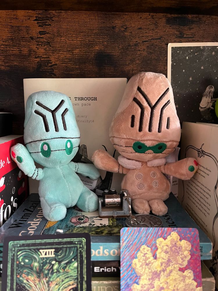 Elgyem and Beheeyem sitting cuties plushies atop a bunch of books. In front are two cards from the Cosma Visions deck. 