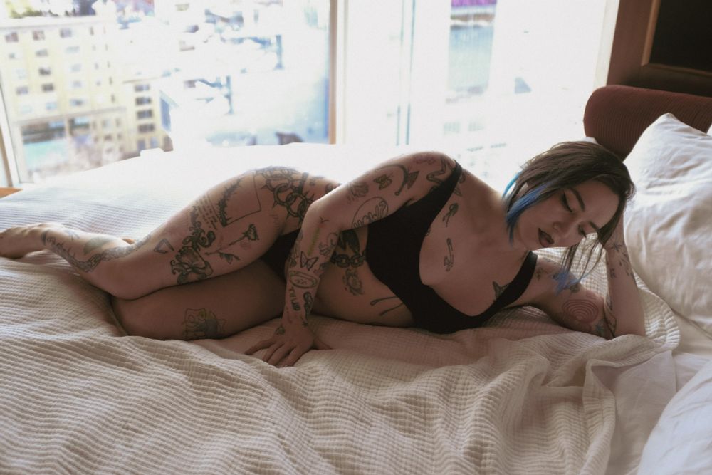 Tattooed girl with dark hair lies on a large round bed with eyes romantically cast downward
