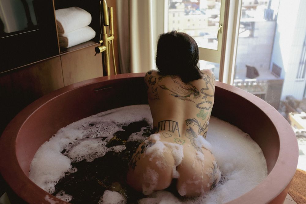 Curvy brunette with tattoos and bubbles on her figure faces away from the camera in a huge round bathtub