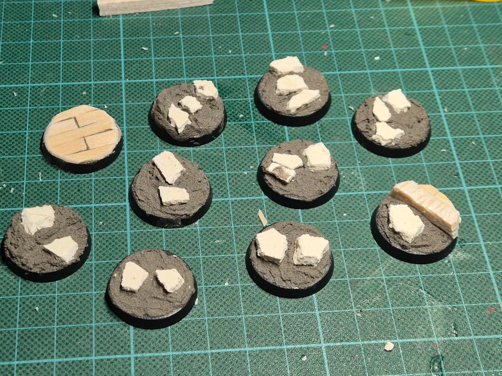 An array of 25mm bases, again on a green cutting board. They have a grey texture paste on them, with some rocks inserted into the pase. One has some floorboards. 