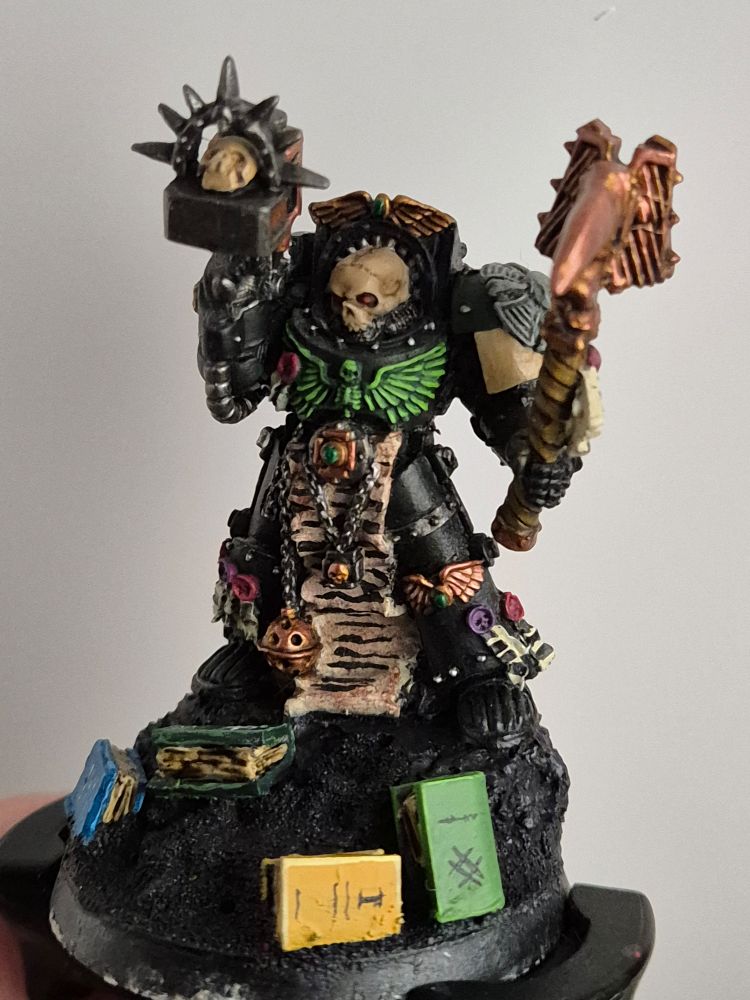 The old terminator chaplain, painted black, golden, and bone. His weapons are golden, his helmet a stark bone with red glowing eyes. A parchment between his legs is scribbled with text - maybe the names of some Fallen he is hunting, eh, searching?
Around the base are some books scattered, half sunken into the mud.