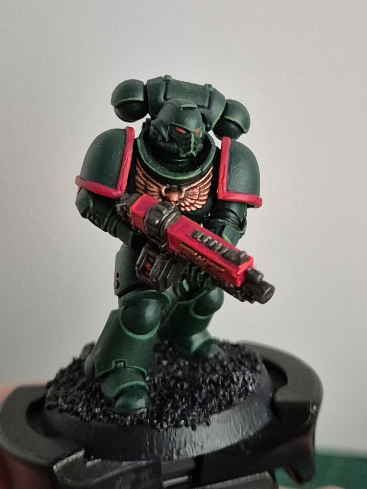 A Primaris Space Marine in the dark green of the Dark Angels. His eyes, bolter and the rim of his pauldrons are a bright red, the decor on his armor bronze.