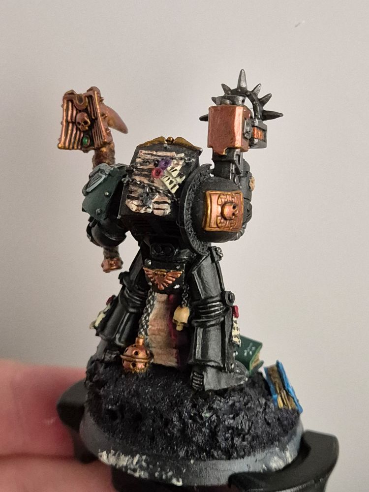 The old terminator chaplain, painted black, golden, and bone. His weapons are golden. He is shown from behind, more golden/bronze accessoires are visible. Another parchment is slung on his back.
