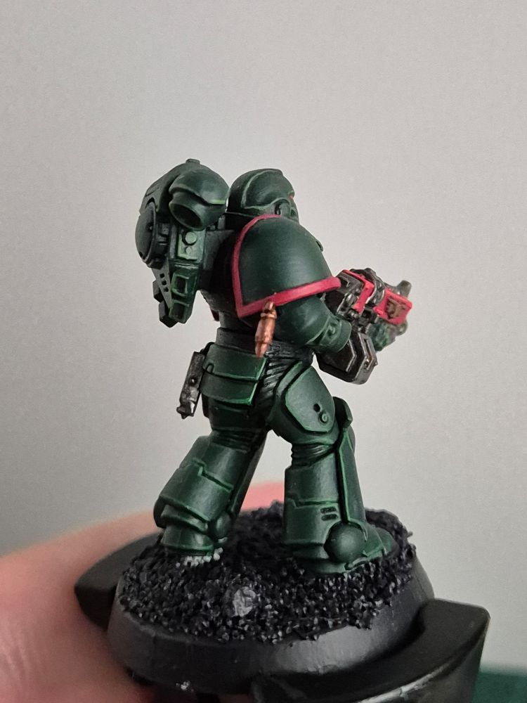 A Primaris Space Marine in the dark green of the Dark Angels. His eyes, bolter and the rim of his pauldrons are a bright red. He is carying a little trinket on his belt and a marksman bullet on his right pauldron.