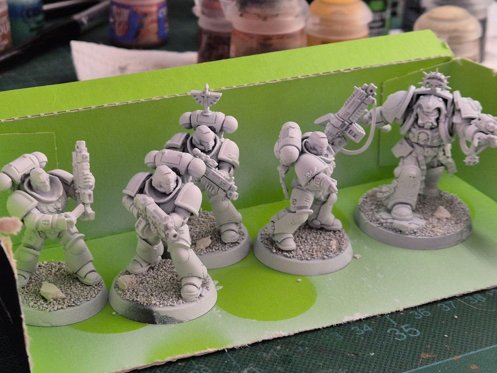 Five Space Marine miniatures on a green cardboard thingy. Four of them are Intercessors: two taking aim, one reloading, one pointing at something. The last one is a Librarian in terminator armour, unleashing the powers of the warp forward... or he is reaching for the last SecretSoda in the cafeteria of the Rock.