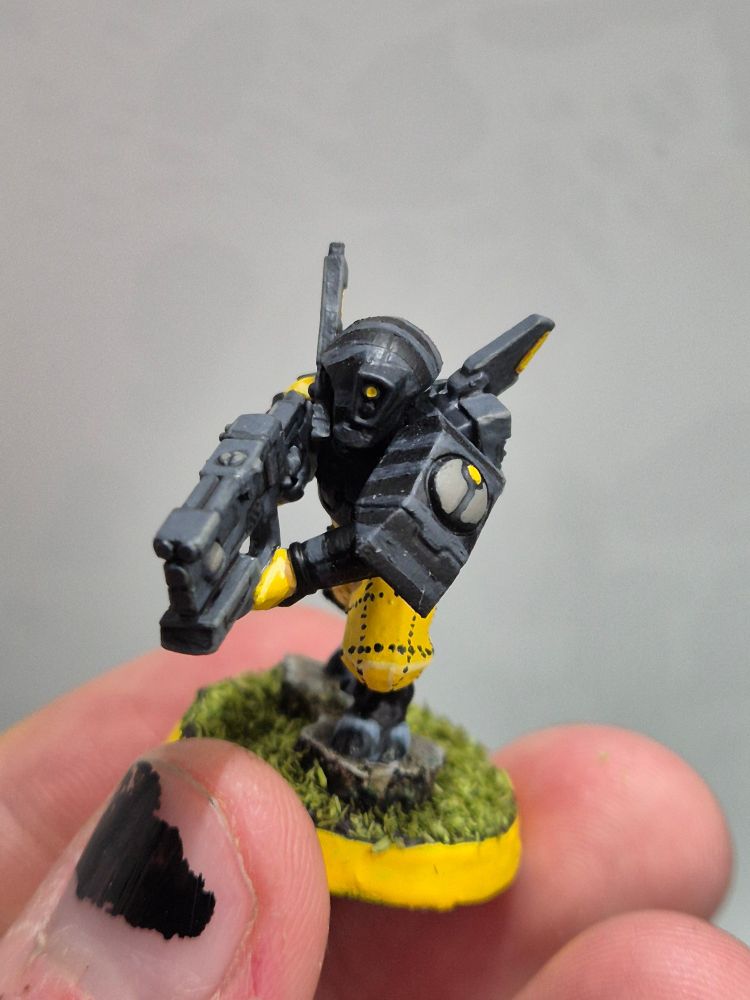 A photo showing a T'au pathfinder. His armor is a dark black, with lighter grey stripes. The sept symbol on his shoulder, taken from a Fire Warrior, is a light grey with a yellow dot on top. He is outfitted with the trappings of a drone controller, carrying the console on his back. His jumpsuit is a bright yellow, with zick-zack dotted lines crosses upon his pants.

He is shown from the front, moving towards the cam.