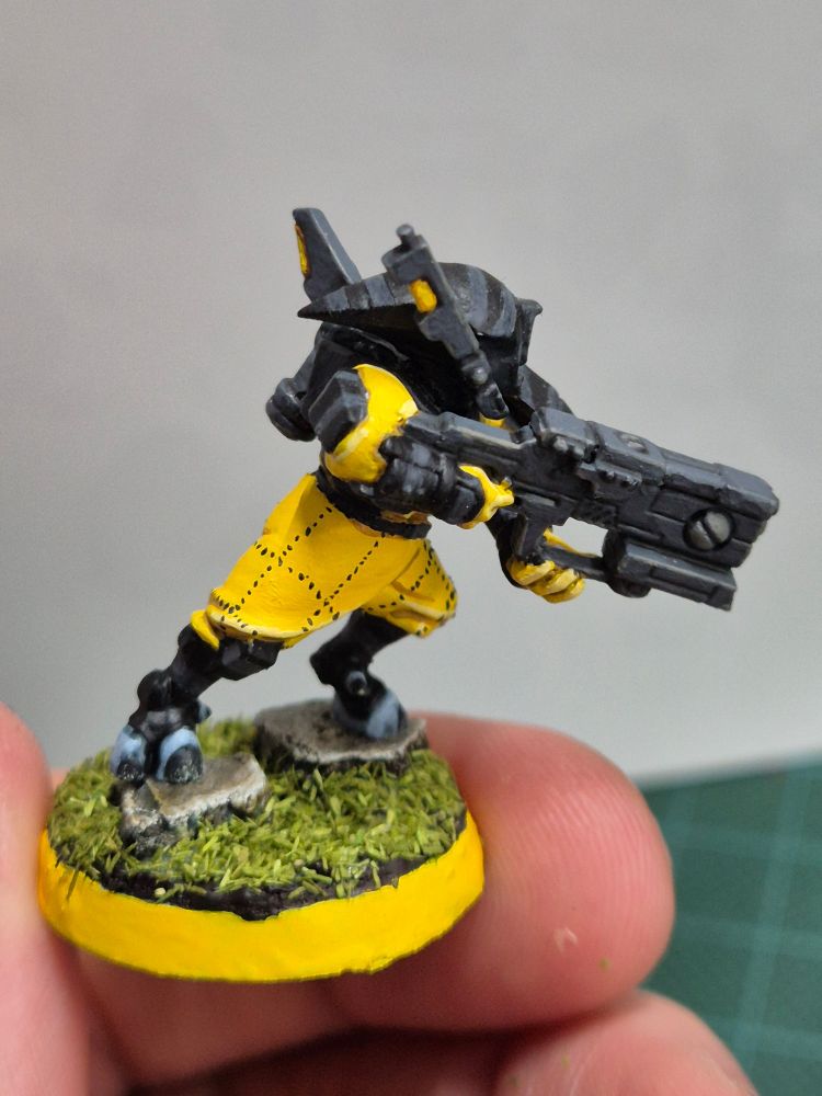 A photo showing a T'au pathfinder. His armor is a dark black, with lighter grey stripes. The sept symbol on his shoulder, taken from a Fire Warrior, is a light grey with a yellow dot on top. He is outfitted with the trappings of a drone controller, carrying the console on his back. His jumpsuit is a bright yellow, with zick-zack dotted lines crosses upon his pants.

He is shown from the other side. He is leaning forward as if in motion, with his carbine on display.