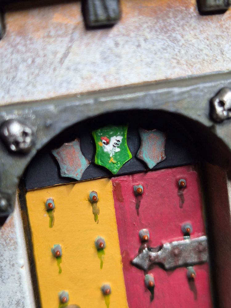 A close up.of the door belonging to the tower of the Mansion of the Empire. Above the door, which is painted in the yellow and red of Talabecland, are three small coat of arm shields - two of them are bronze and heavily patina's, but the third is coloured a verdant green. Upon the green, a very simple and tine chicken is painted. It's something, I guess.